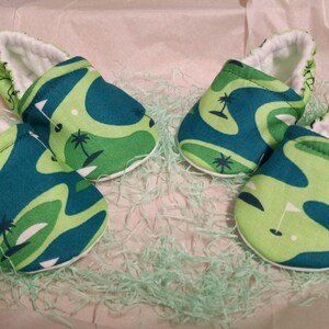 Puede incluir: Cuatro zapatos de beb&eacute; con un dise&ntilde;o de campo de golf. Los zapatos presentan un patr&oacute;n de formas verdes y turquesas con palmeras y banderas de golf. El interior de los zapatos es blanco y est&aacute;n colocados sobre papel verde.