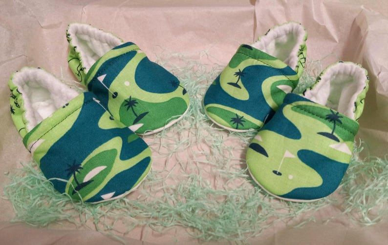 Puede incluir: Cuatro zapatos de beb&eacute; con un dise&ntilde;o de campo de golf. Los zapatos presentan un patr&oacute;n de formas verdes y turquesas con palmeras y banderas de golf. El interior de los zapatos es blanco y est&aacute;n colocados sobre papel verde.