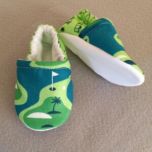 Puede incluir: Zapatos de beb&eacute; con dise&ntilde;o de campo de golf. Los zapatos presentan un patr&oacute;n verde y azul, suelas blancas e interiores blancos. El dise&ntilde;o incluye una bandera de golf, una palmera y un campo de golf. Miden unos 10 cm.