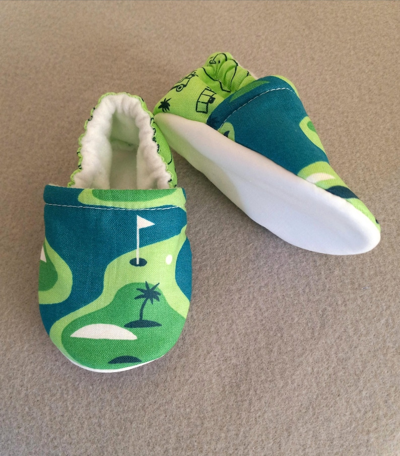 Puede incluir: Zapatos de beb&eacute; con dise&ntilde;o de campo de golf. Los zapatos presentan un patr&oacute;n verde y azul, suelas blancas e interiores blancos. El dise&ntilde;o incluye una bandera de golf, una palmera y un campo de golf. Miden unos 10 cm.