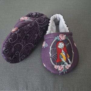 May include: Two purple baby booties. One bootie has a floral pattern, the other features a Sally from The Nightmare Before Christmas design. The booties have white lining and are designed for comfort.