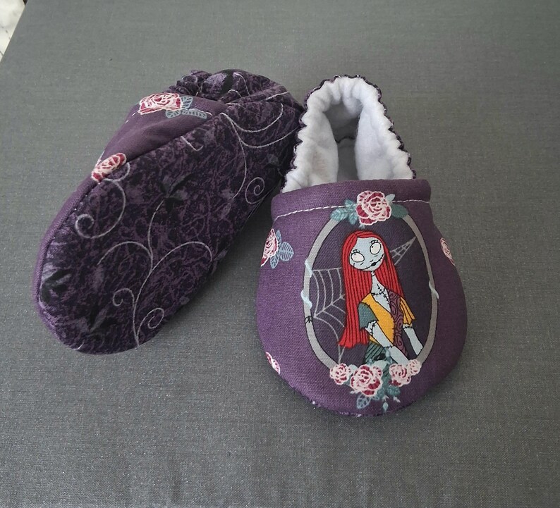 May include: Two purple baby booties. One bootie has a floral pattern, the other features a Sally from The Nightmare Before Christmas design. The booties have white lining and are designed for comfort.