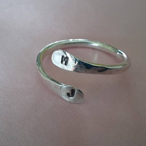 May include: A silver ring with a hammered finish. The ring is wrapped around the finger and has the initials "M" and "J" engraved on it.