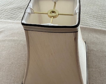 May include: A white lampshade with a black trim and a gold metal harp. The lampshade is square at the top and bottom and has a slight bell shape.
