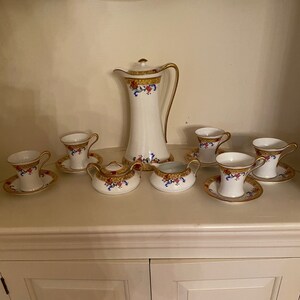 Peut inclure: Un service à thé en porcelaine blanche avec des bordures dorées et des motifs floraux. L'ensemble comprend une théière, un pot à crème, un sucrier et six tasses et soucoupes.