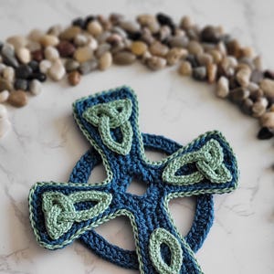 Op de afbeelding: Een gehaakt Keltisch knoopkruis in blauw en groen. Het kruis is omringd door kleine stenen.