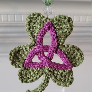 May include: A green crocheted shamrock with a pink Celtic knot design. The shamrock is hanging from a string with clear and green beads.