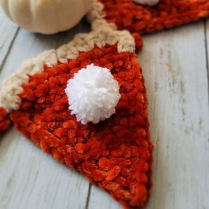 May include: A garland of crocheted pumpkin pie slices with white pom-poms. The garland is on a white wooden surface with a white pumpkin in the foreground.