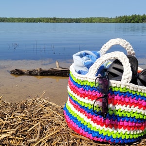 May include: A colorful striped crochet beach bag with white handles. The bag is filled with a blue towel, sunglasses, and flip-flops. The bag is sitting on a sandy beach.