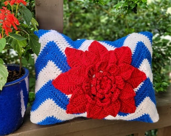 May include: A handmade crochet pillow with a red flower applique. The pillow features a blue and white chevron pattern. A potted plant with red flowers sits to the left of the pillow.