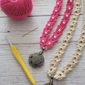 May include: Two crocheted necklaces with metal charms. The pink necklace has white beads and the beige necklace has a flower design. Both charms are round and have the word "Clover" on them.