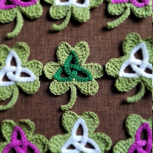 May include: A collection of crocheted shamrock ornaments with different colored Celtic knot designs. The shamrocks are green with white, pink, and green knots.
