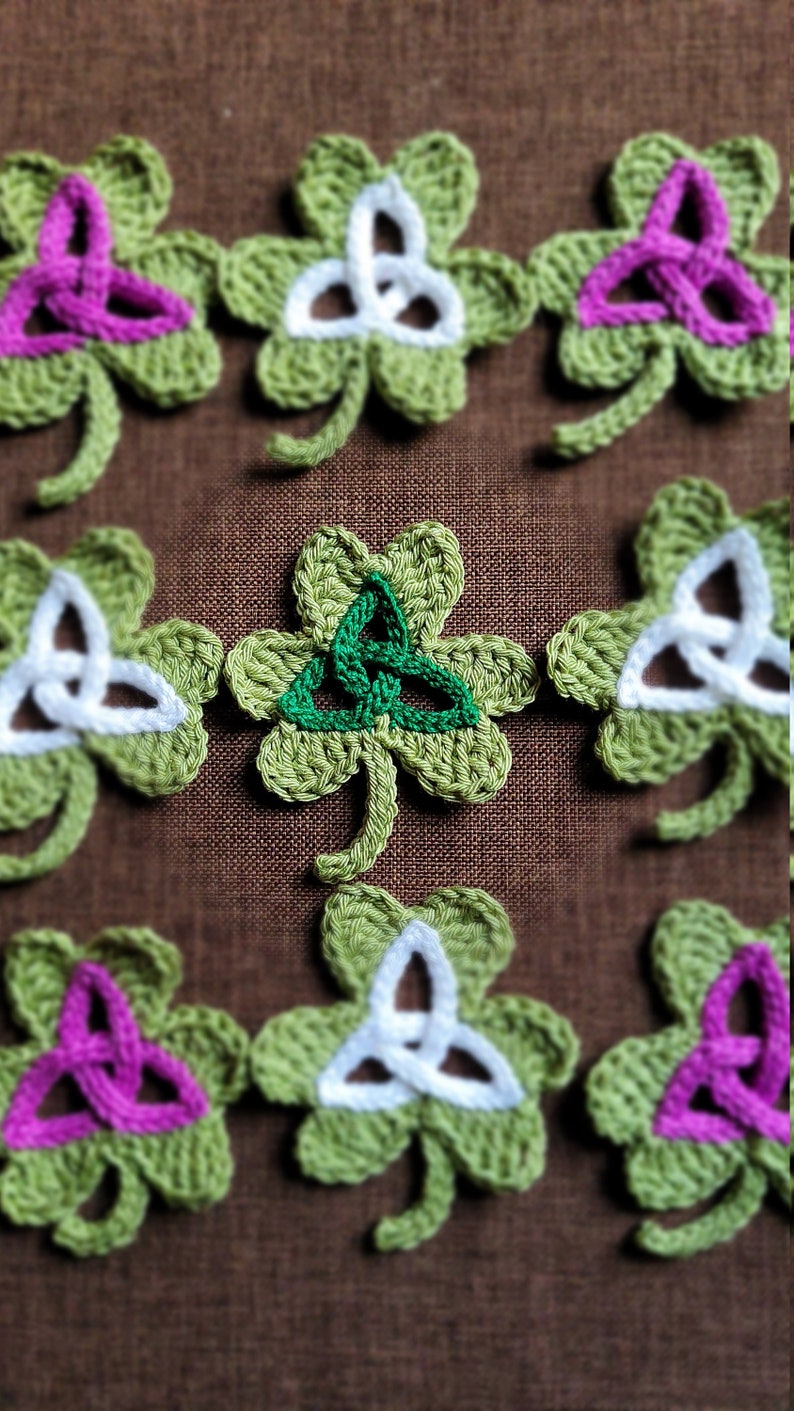 May include: A collection of crocheted shamrock ornaments with different colored Celtic knot designs. The shamrocks are green with white, pink, and green knots.