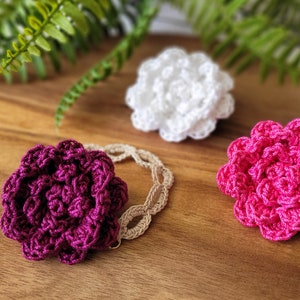 May include: Three crocheted flowers in different colours: pink, white and purple. The flowers are displayed on a wooden surface.