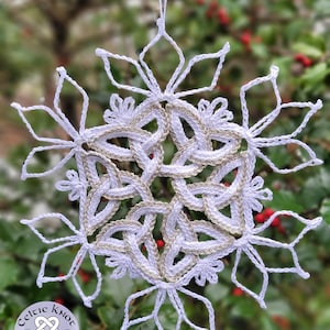 以下が含まれることがあります： 金色のケルト結び目のデザインが施された白いクロシェの雪の結晶のオーナメント。オーナメントは赤い実のついた枝にぶら下がっています。