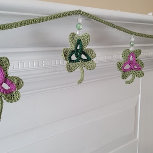 May include: A green crocheted garland with shamrock charms featuring a Celtic knot design. The shamrocks are decorated with pink, green, and white yarn. The garland is strung with clear beads.