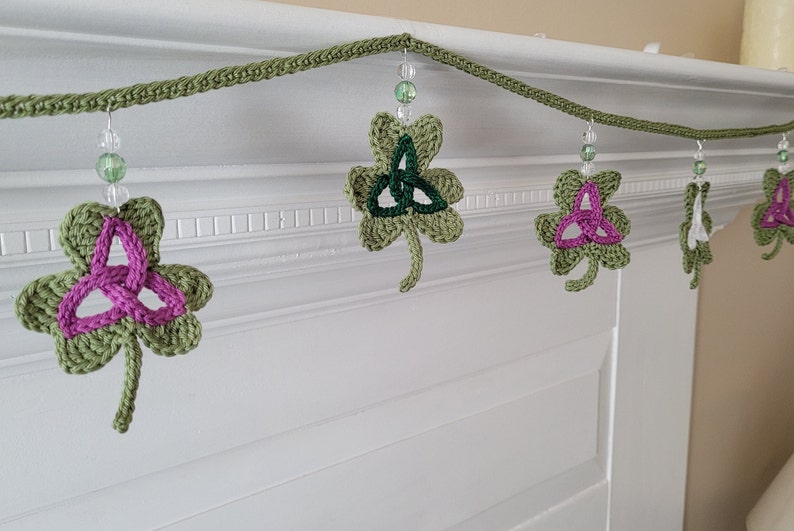 May include: A green crocheted garland with shamrock charms featuring a Celtic knot design. The shamrocks are decorated with pink, green, and white yarn. The garland is strung with clear beads.