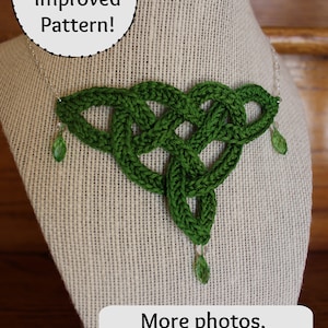 Pode incluir: Um colar de nó celta de crochê verde com detalhes de cristal. O colar é exibido em um manequim coberto de tecido. O texto "Updated & Improved Pattern!" está em um círculo acima do colar. O texto "More photos, better diagrams & a video tutorial" está abaixo do colar. Um rosto sorridente está à direita do texto.