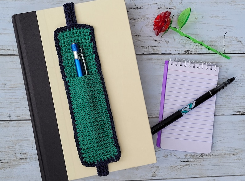 Op de afbeelding: Een groene en marineblauwe gehaakte bladwijzer met een penhouder. De bladwijzer is bevestigd aan een boek met een gele kaft. Een klein notitieboekje en een pen zijn ook zichtbaar.