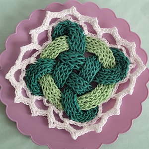 May include: A green and white crocheted doily with a woven knot design in the center. The doily is on a purple background.