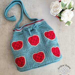 May include: A blue crocheted tote bag with a red heart design. The bag has a long strap and a drawstring closure. The bag is decorated with five red hearts, each with a white outline. The bag is made by Celtic Knot Crochet.