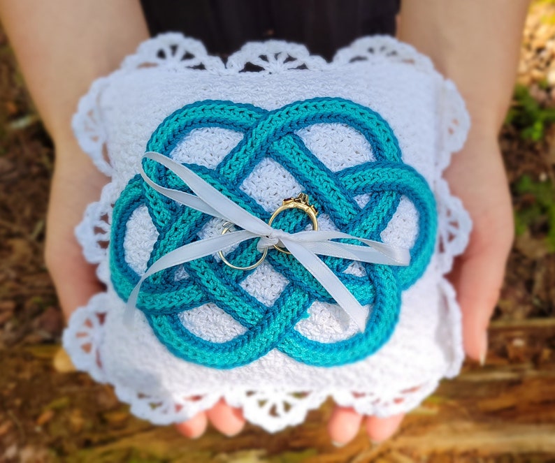 May include: White crocheted cushion with a blue Celtic knot design, featuring two gold rings tied with a white satin ribbon.