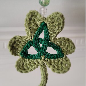 May include: A green crocheted shamrock with a green Celtic knot in the center. The shamrock is hanging from a string of beads.