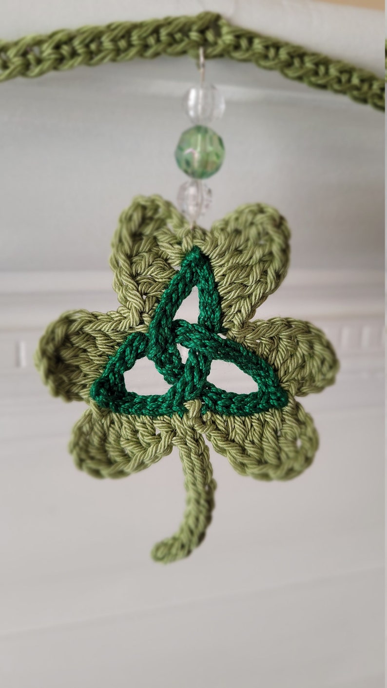 May include: A green crocheted shamrock with a green Celtic knot in the center. The shamrock is hanging from a string of beads.