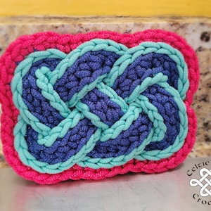 May include: A crocheted Celtic knot in pink, teal, and blue. The knot is in the shape of a rectangle and has a raised design. The knot is on a granite countertop.
