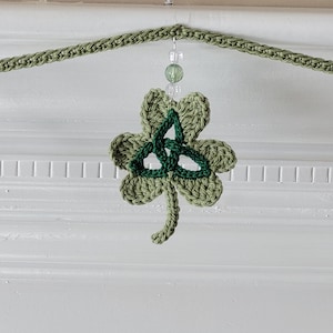 May include: A green crocheted shamrock garland with pink and green Celtic knot designs. The garland is strung with clear beads and is perfect for St. Patrick's Day decor.