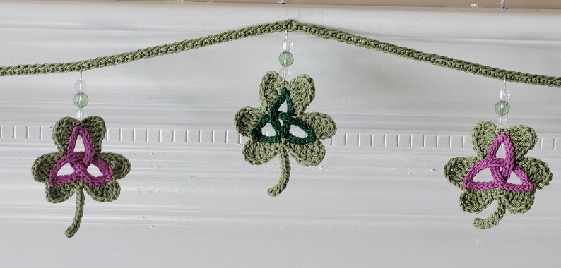 May include: A green crocheted shamrock garland with pink and green Celtic knot designs. The garland is strung with clear beads and is perfect for St. Patrick's Day decor.