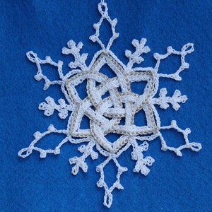 Pode incluir: Um enfeite de floco de neve branco de crochê com um centro metálico prateado. O floco de neve está sobre um fundo azul.