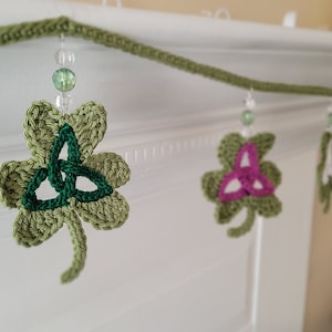 May include: A green crocheted shamrock garland with green and white beads hanging from it. The garland is decorated with a green shamrock with a green Celtic knot in the center and a pink shamrock with a pink Celtic knot in the center.