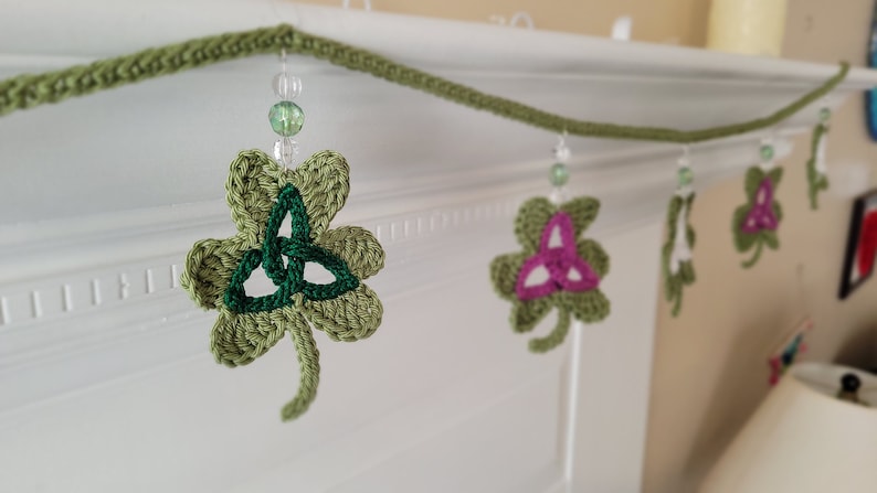 May include: A green crocheted shamrock garland with green and white beads hanging from it. The garland is decorated with a green shamrock with a green Celtic knot in the center and a pink shamrock with a pink Celtic knot in the center.