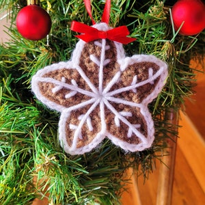 May include: A brown crocheted star-shaped ornament with a white snowflake design hangs on a green Christmas tree with red ornaments.