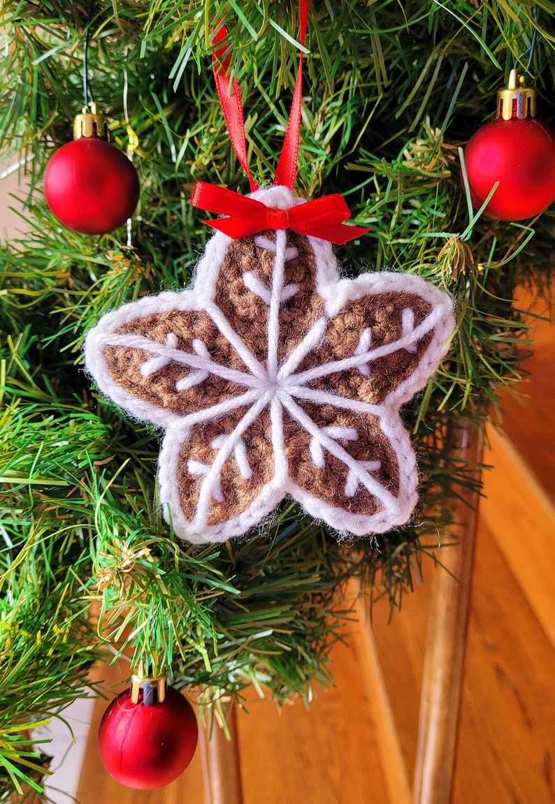 May include: A brown crocheted star-shaped ornament with a white snowflake design hangs on a green Christmas tree with red ornaments.