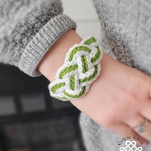 Könnte beinhalten: Ein gehäkeltes Armband in Grün und Weiß mit einem keltischen Knotendesign. Das Armband wird am Handgelenk getragen.