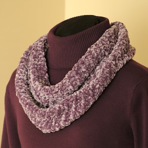 Può includere: Una sciarpa a infinito in peluche color lavanda è drappeggiata attorno a un dolcevita viola scuro. La sciarpa ha un aspetto morbido e strutturato ed è realizzata in un materiale spesso e vellutato. Il dolcevita è di colore pieno, creando un contrasto con la trama della sciarpa.