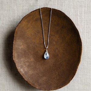 May include: A silver necklace with a teardrop-shaped, pale blue gemstone pendant. The pendant hangs from a delicate chain. The necklace rests on a brown, round plate with a textured surface, set against a neutral background.