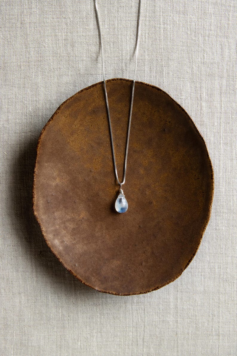 May include: A silver necklace with a teardrop-shaped, pale blue gemstone pendant. The pendant hangs from a delicate chain. The necklace rests on a brown, round plate with a textured surface, set against a neutral background.