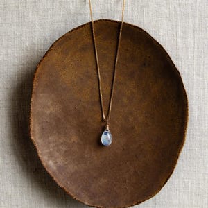 May include: A gold-colored necklace with a teardrop-shaped, light blue gemstone pendant. The necklace rests on a rustic, brown, circular plate. The plate is set against a neutral, textured background.