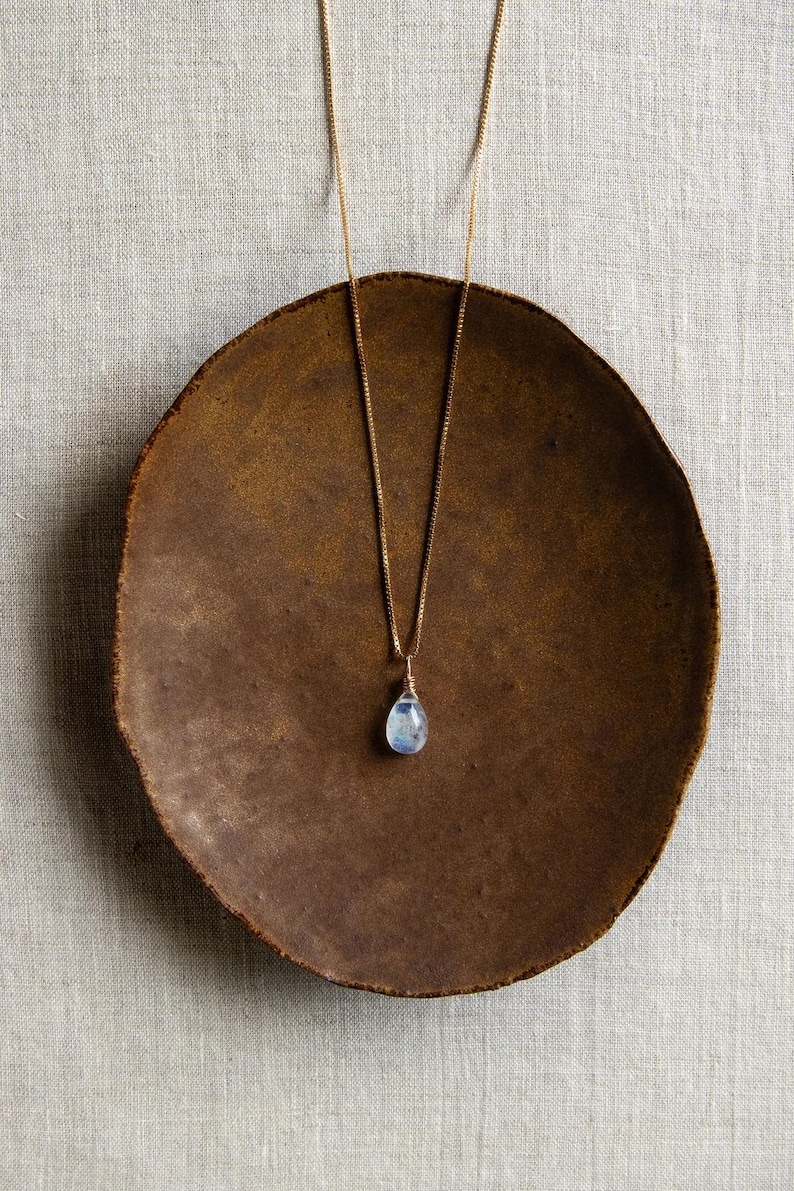 May include: A gold-colored necklace with a teardrop-shaped, light blue gemstone pendant. The necklace rests on a rustic, brown, circular plate. The plate is set against a neutral, textured background.