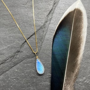May include: A gold chain necklace with a teardrop-shaped moonstone pendant. The pendant is wrapped in gold wire and has a iridescent blue and white color.