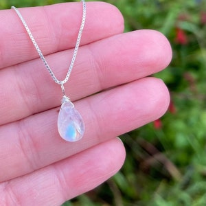 May include: A delicate silver necklace featuring a pear-shaped moonstone pendant. The iridescent gemstone displays a subtle play of blue and white hues. The necklace is held against a blurred green and red background.