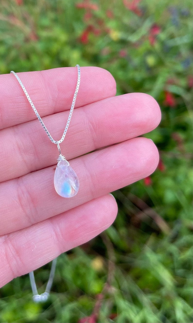 May include: A delicate silver necklace featuring a pear-shaped moonstone pendant. The iridescent gemstone displays a subtle play of blue and white hues. The necklace is held against a blurred green and red background.