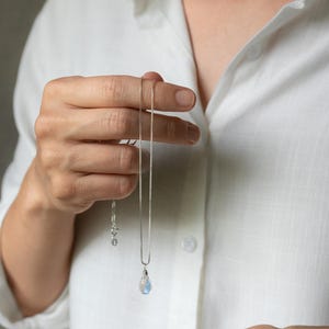 May include: A silver necklace with a teardrop-shaped pendant. The pendant has a blue and white iridescent appearance. The necklace is held against a white shirt, showcasing the jewelry's delicate design. The chain is a fine box chain.