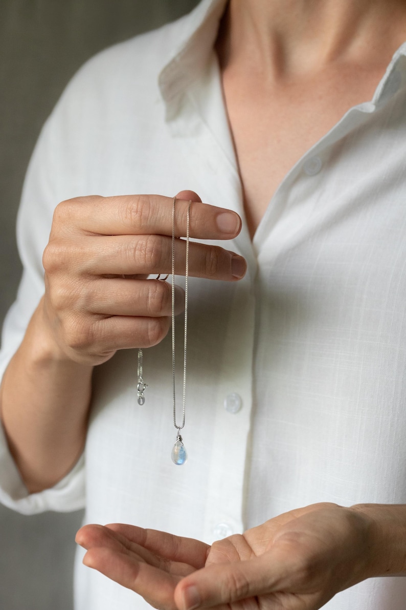 May include: A silver necklace with a teardrop-shaped pendant. The pendant has a blue and white iridescent appearance. The necklace is held against a white shirt, showcasing the jewelry's delicate design. The chain is a fine box chain.