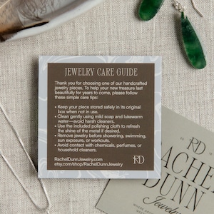 May include: An image featuring a silver necklace with a teardrop-shaped pendant, green jade earrings, and a jewelry care guide. The guide provides care tips. A business card with the text "Rachel Dunn Jewelry" is also visible.