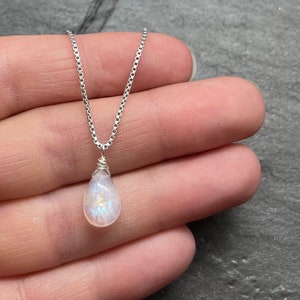 May include: A delicate necklace featuring a pear-shaped, iridescent moonstone pendant. The pendant is wire-wrapped and suspended from a fine, silver chain. The necklace is displayed against a neutral background.