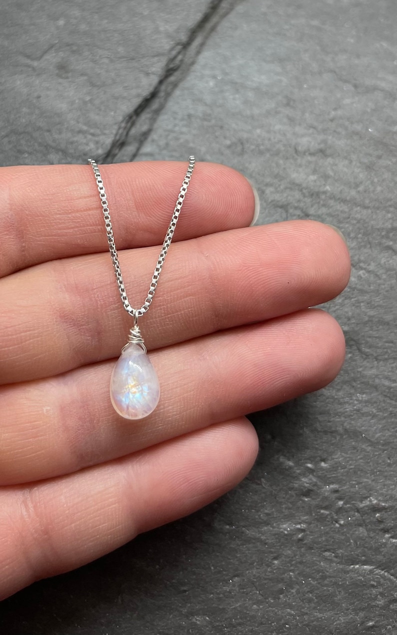 May include: A delicate necklace featuring a pear-shaped, iridescent moonstone pendant. The pendant is wire-wrapped and suspended from a fine, silver chain. The necklace is displayed against a neutral background.